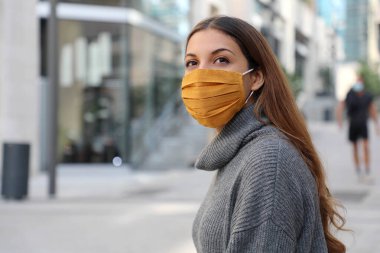 Koruyucu maskeli genç bir kadın modern şehir sokaklarına bakıyor.