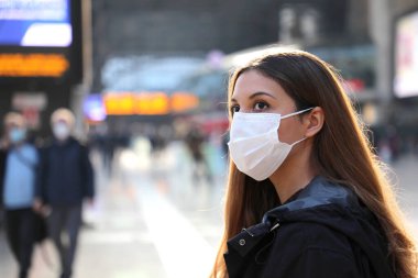 Havaalanında cerrahi maske takan gezgin kadına yakın çekim. Endişeli genç bir kadın varış saatlerine bakıyor. Uçağı için bilgi bekliyor..