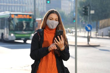 Koruyucu maskeli genç bir iş kadını akıllı telefon uygulamasından sohbet mesajlarını kontrol ediyor ve sabah sokakta otobüs bekliyor.