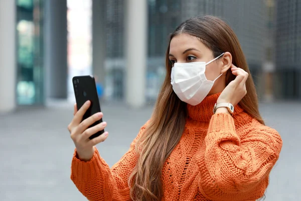 Video calling and Social Distancing. Beautiful young woman in city street wearing protective mask using mobile phone to make video calling or video conference outdoor.