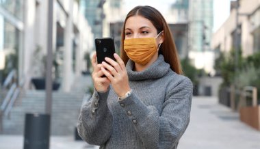 Koruyucu maske takmış ve akıllı telefonla mesajlaşan genç bir kadının portresi.