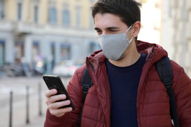 Koruyucu maskeli mutlu genç adam akıllı telefonunu sokakta görüyor. Yüzü maskeli öğrenci dışarıda cep telefonu kullanıyor..