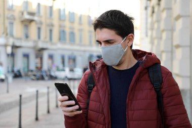 Beyaz adam sokakta yürürken cep telefonu kullanarak yüz maskesi takıyor.