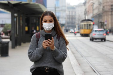 Koruyucu maskeli güzel bir kadın şehir caddesinde akıllı telefonlardan online taşıma bileti alıp parasını ödüyor.