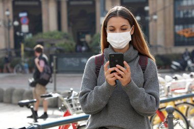Akıllı telefon kullanan ve şehir merkezinde sergilenen koruyucu maskeli genç bir kadın.