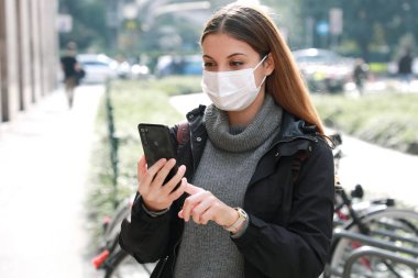 Akıllı telefon kullanan ve şehir merkezinde sergilenen koruyucu maskeli genç bir kadın.