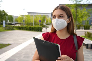Portrait of beautiful girl with FFP2 KN95 face mask holding folders looking to the side outdoors clipart