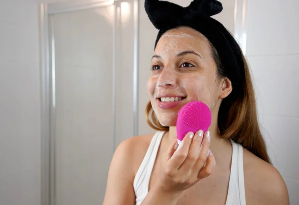 Close up of smiling girl cleaning washing skin with silicon brush ultrasonic massage in the bathroom