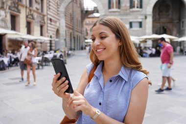 Nice girl scrolling on her mobile phone outdoors. Smiling attractive stylish woman typing on smartphone in Verona square, Italy.
