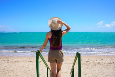 Cennet kumsalı konsepti. Gezgin kadının arka görüntüsü merdivenlerden aşağı, plajın kristal suyuna doğru iniyor..