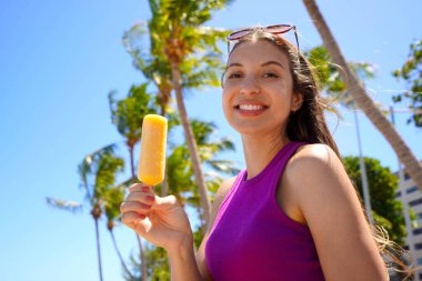Sahilde buzlu dondurma yiyen gülümseyen genç bir kadın. Arka planda palmiye ağaçları var. Kameraya bak..