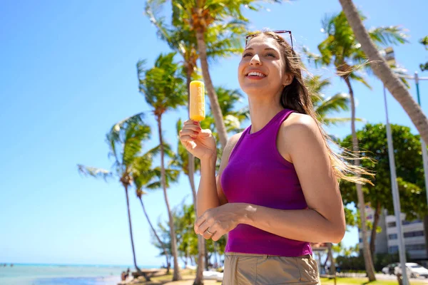 Plajda buzlu dondurma yiyen genç bir kadın. Arka planda palmiye ağaçları var.