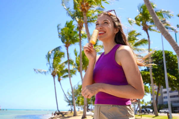 Sahilde buzlu dondurma yiyen güzel bir kadın. Arka planda palmiye ağaçları var.