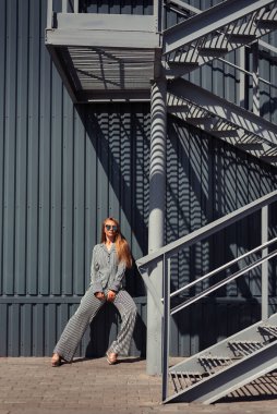 Çizgili gömlekli ve pantolonlu bir kız demir merdivenin yanında duruyor. Sokak vuruşu güzel kız.