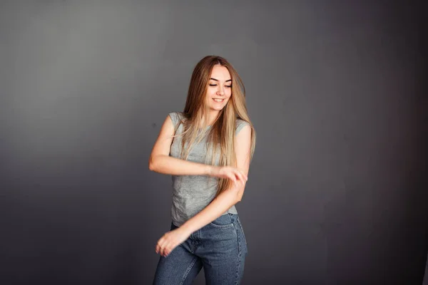 金髪の女の子は 灰色の背景にプレーングレーのTシャツとジーンズに手を上げて踊る — ストック写真