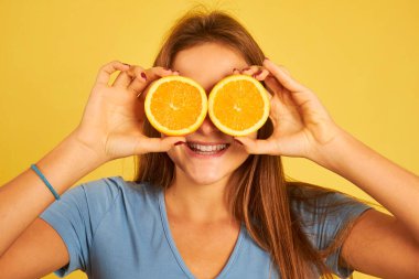 Sarı arka planda portakalları gözlük olarak kullanan mutlu genç bir kadın.