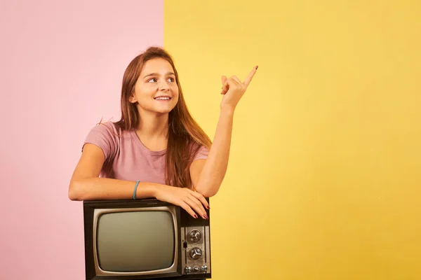 年轻女子手握复古电视机 坐在黄色和粉色的背景下 手指指向旁边 — 图库照片