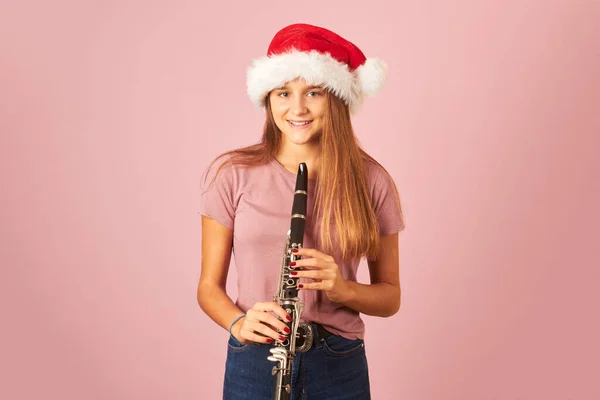 young performer woman playing clarinet and wearing Santa Claus hat on a pink background