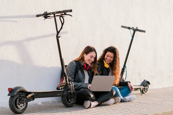 Két többnemzetiségű női barát ül az utcán, elektromos robogókkal laptoppal Jogdíjmentes Stock Fotók