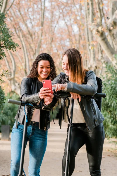 Zwei multiethnische Freundinnen auf Elektrorollern mit Smartphone im Freien Stockbild