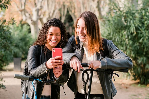 Twee multi-etnische vrouwelijke vrienden op elektrische scooters met behulp van smartphone buiten Stockfoto