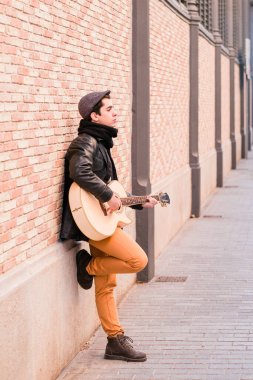 Sokak müzisyeni akustik gitar çalıyor. Paltolu ve şapkalı genç bir delikanlı.