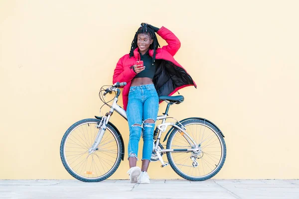 Boldog afro-amerikai nő visel piros kabát kerékpárral az utcán sárga háttér. — Stock Fotó