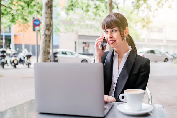 Porträt einer schönen Geschäftsfrau mit Laptop, die im Café sitzt und ihr Smartphone benutzt lizenzfreie Stockbilder