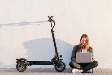 Elektrikli scooterlı genç bir kızın portresi. Kırmızı kulaklıklı beyaz bir duvarın üzerinde dizüstü bilgisayar kullanan bir kadın. Kentsel yaşam tarzı ve ekolojik ulaşım kavramları