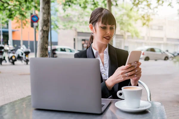Портрет красивой деловой женщины с ноутбуком, сидящей в кофейне на открытом воздухе и использующей смартфон — стоковое фото