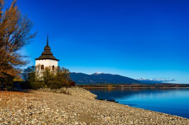 Güneşli bir sonbahar gününde Liptovska Mara su deposunun kıyısında tarihi bir beyaz kilise kulesi. Arka planda, Batı ve Yüksek Tatra 'nın karlı zirveleri açık mavi bir gökyüzünün altında görülebilir. Slovakya, Liptov bölgesi.