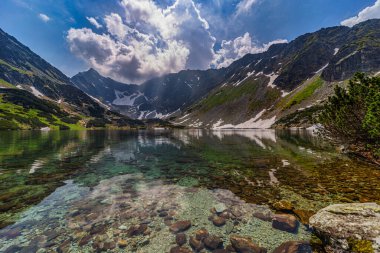 Slovakya 'daki High Tatras Ulusal Parkı' ndaki buzul gölü Nizne Temnosmrecinske Pleso 'nun çarpıcı manzarası. Görüntü, altında kayalar olan şeffaf suyu, sivri dağ tepelerini çevreleyen kayaları ve sakin surfeye yansıyan dramatik gökyüzünü içeriyor.