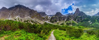 Yeşil Göl 'ün kıyısında (Zelene pleso) bir yürüyüş yolunun dramatik bir manzarası. Şiddetli gri fırtına bulutlarıyla kaplı, kar yamaçları olan devasa granit tepeler. Tatra Ulusal Parkı, Slovakya.
