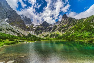 Slovakya 'nın High Tatras kentindeki Zelene pleso' nun (Yeşil Göl) nefes kesen geniş manzarası. Görüntüde ikonik dağ kulübesi, kristal berrak su, kayalık tabanı ve dinamik bir yaz gökyüzü altında pürüzsüz granit tepeler yer alıyor..
