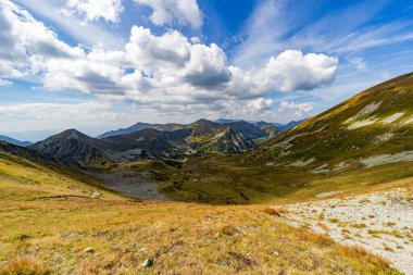 Dar bir kayalık yol bir dağ zirvesine doğru dik bir çimenli yamaçtan geçer. High Tatras Ulusal Parkı 'ndaki Hladke Sedlo' dan manzara, bulutlu mavi gökyüzünün altında vadide turkuaz bir göl yer alıyor.
