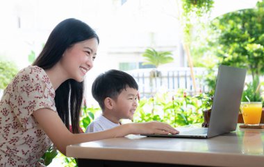 Genç Asyalı anne ve oğlu evde birlikte çalışmak ve öğrenmek için dizüstü bilgisayar kullanıyor, uzaktan eğitim, öğretmen veya anne desteği, eğitim konsepti ile e-öğrenim için kulaklık takıyor..
