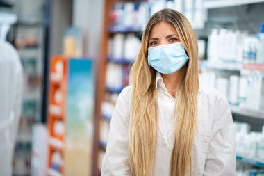 Gülümseyen genç sarışın eczacı eczanesinin içinde Coronavirus maskesi takıyor.