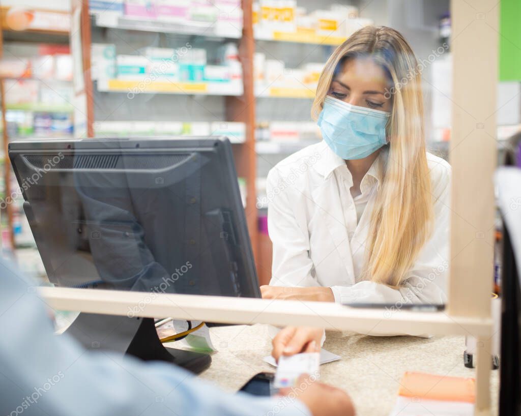 Smiling young blonde pharmacist wearing covid coronavirus mask inside her pharmacy
