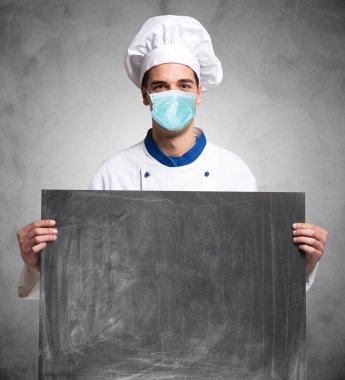 Maskeli aşçı ya da aşçı covid koronavirüs salgını, restoranlar ve yemek konsepti sırasında karatahta tutuyor.