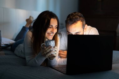 Geceleri yatarken dizüstü bilgisayar kullanan neşeli genç bir çiftin portresi.