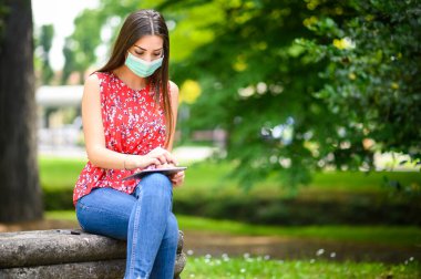 Genç bir kadın koronavirüs zamanlarında parktaki bankta dijital tablet kullanıyor.