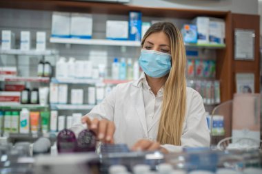 Coronavirus covid maskesi takıp eczanedeki envanteri kontrol eden genç bir kadın eczacı.