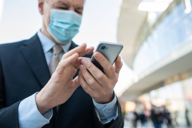 Covid Coronavirus konsepti, maskeli yaşlı iş adamı akıllı telefonunu şehirde kullanıyor.