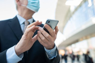 Covid konsepti, maskeli yaşlı iş adamı akıllı telefonunu açık havada bir şehirde kullanıyor.