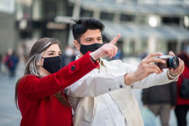 Covid ya da Coronavirus maskeli mutlu turistler bir şehirde birlikte yürüyor ve fotoğraf çekmek için ilginç bir nokta işaret ediyor.