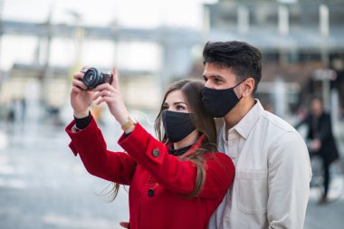Covid ya da Coronavirus maskeli mutlu turistler şehirde birlikte yürüyüp ilginç bir noktayı fotoğraflıyorlar.