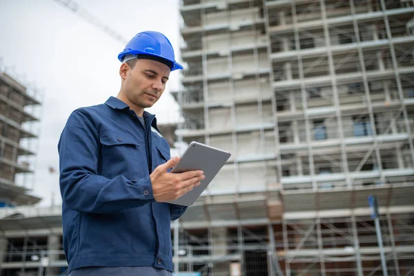 Takım elbiseli ve kasklı genç mühendis inşaat alanının önünde tablet bilgisayarda çalışıyor.