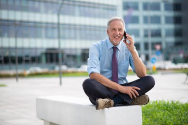 Bir kentteki bankta otururken cep telefonuyla gülümseyen kıdemli bir işadamının portresi.