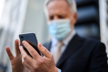 Üst düzey yönetici Coronavirus salgınından korunmak için maske takarken akıllı telefonunu açık havada kullanır.