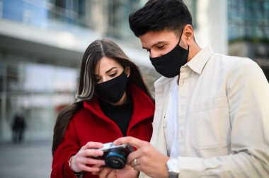 Covid ya da Coronavirus maskeli mutlu turistler bir şehirde birlikte yürüyüp birlikte çekilen fotoğraflara bakıyorlar.